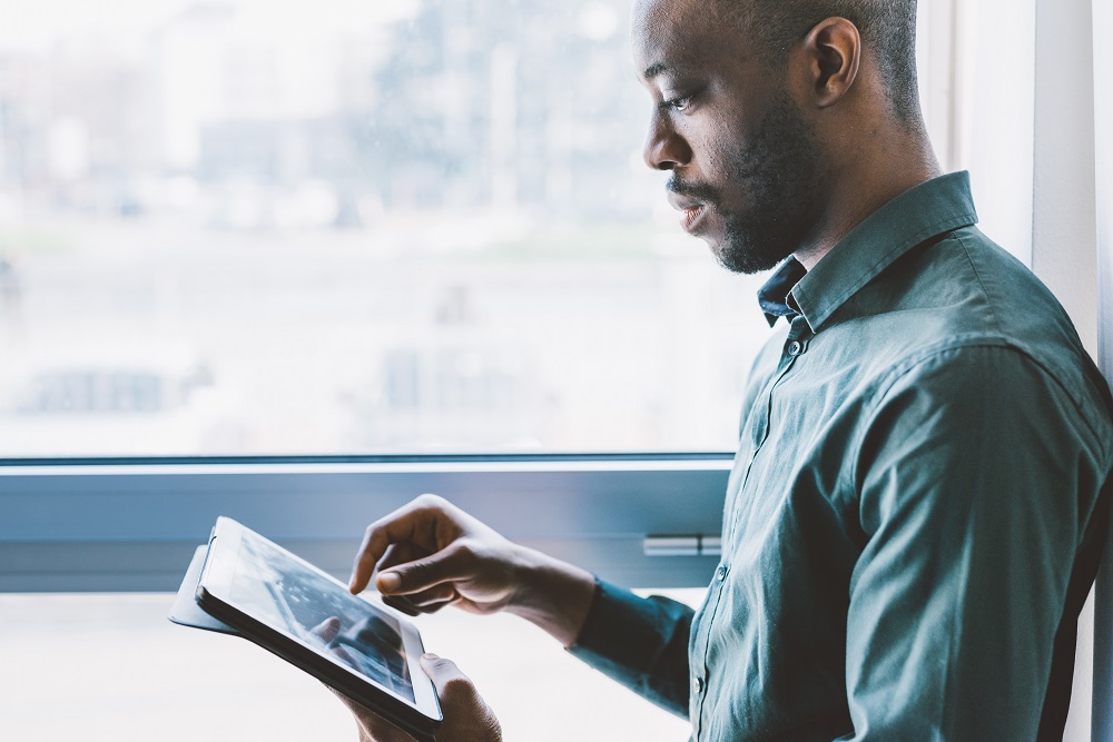 Half length of young handsome afro businessman near a window holding a tablet, looking down and tapping the screen - business, working, job concept