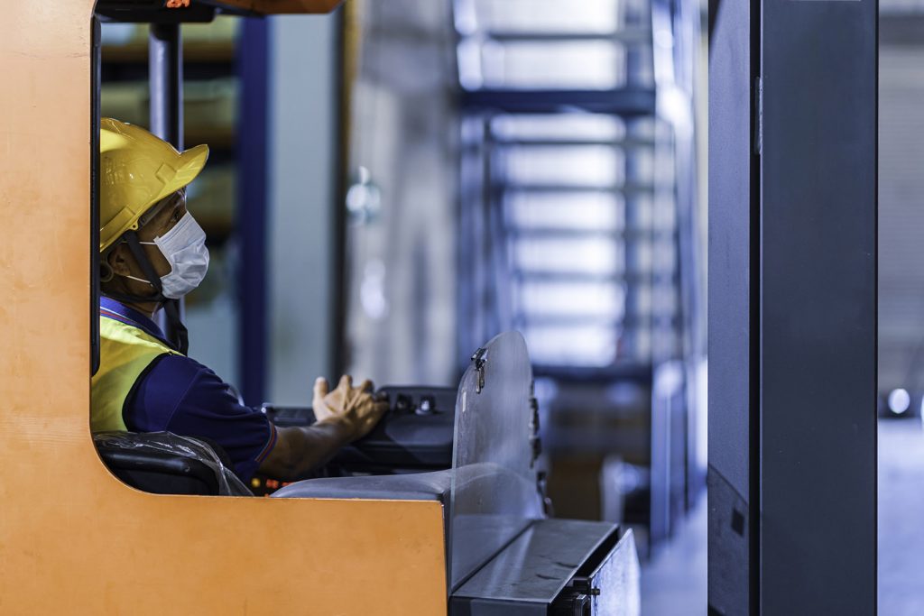 Asian foreman in uniform with helmet and protective face mask drive forklift in warehouse cargo with copy space. Concept of new normal work in logistic industry, factory after Covid 19 pandemic