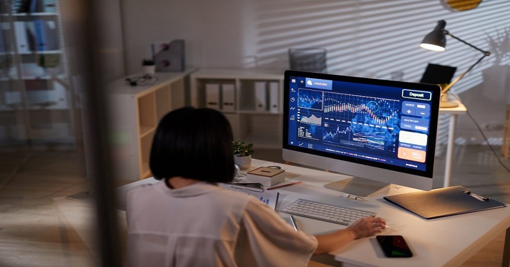 Rear view of young analyst sitting at the table in front of computer monitor with financial charts and analyzing it at office